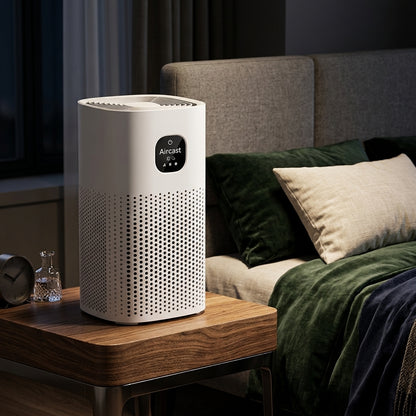 A white Aircast air purifier sits on a wooden bedside table in a modern bedroom setting, next to a bed with green and beige bedding.