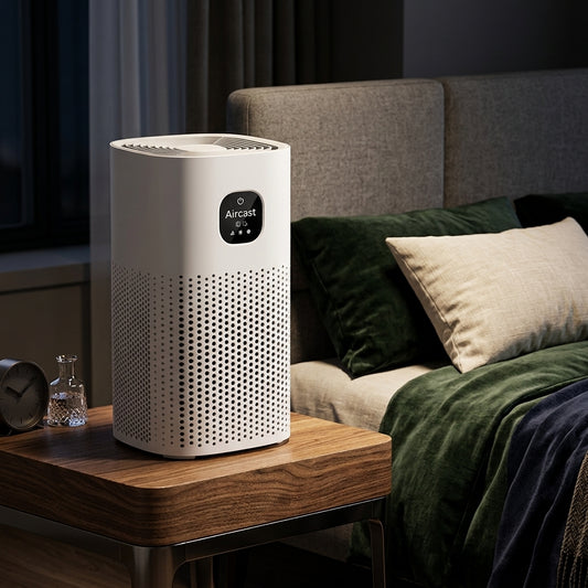 A white Aircast air purifier sits on a wooden bedside table in a modern bedroom setting, next to a bed with green and beige bedding.